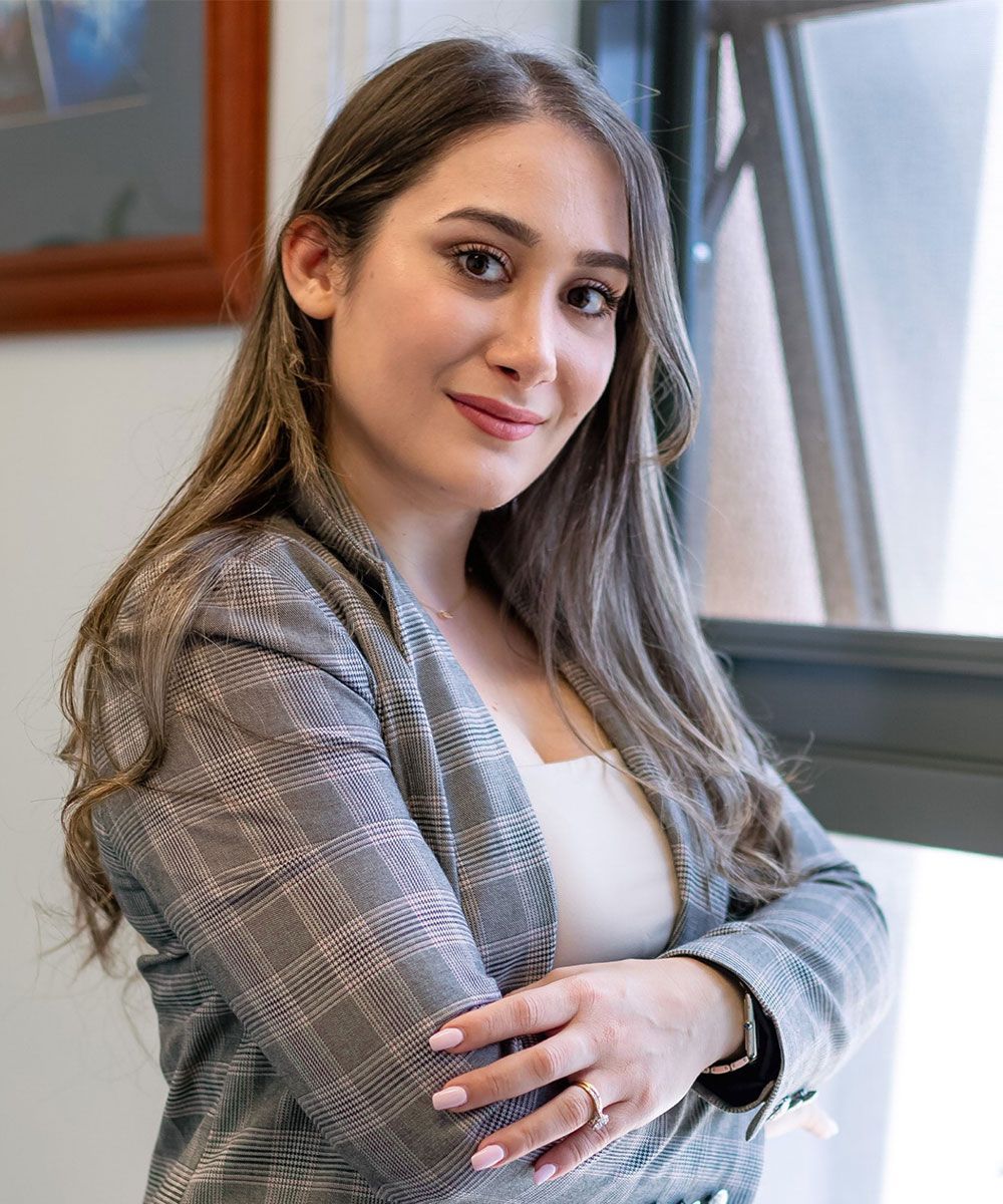 Woman in a plaid blazer, arms crossed, smiling. Leaning against a window with natural light.