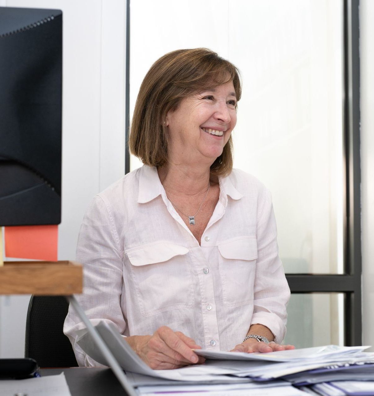 Woman smiles, seated at a desk with papers. Light pink blouse, short brown hair, office setting.