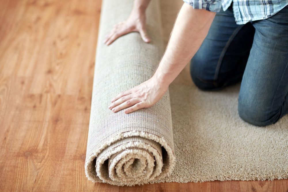 A person kneeling on a wooden floor, unrolling a beige carpet.
