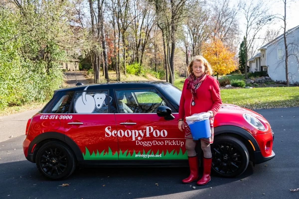 Woman beside red-and-black ScoopyPoo grooming van on a tree-lined street