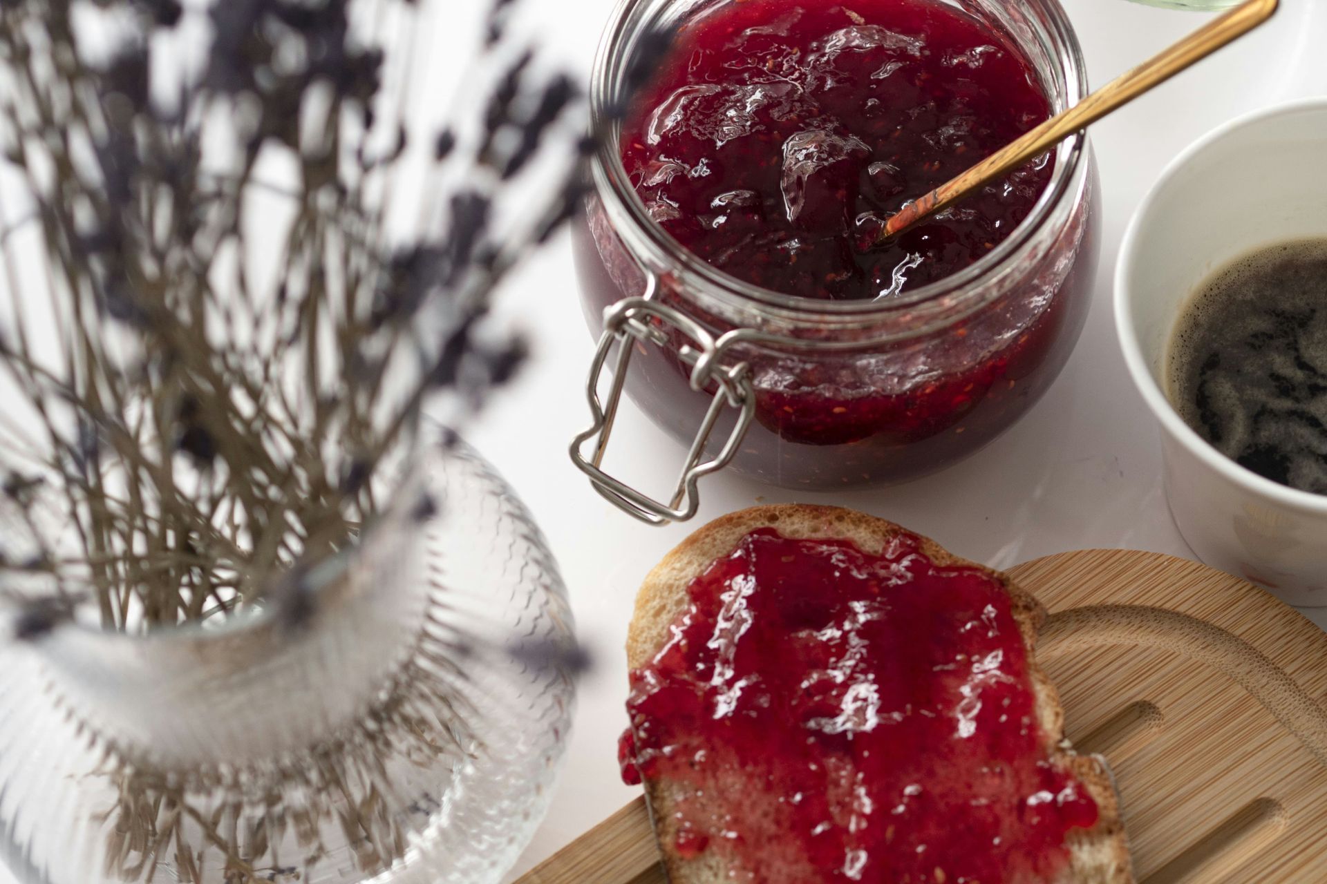 Ein Glas rote Marmelade mit einem goldenen Löffel, eine mit Marmelade bestrichene Scheibe Toast und getrockneter Lavendel in einer Vase auf einem weißen Tisch.