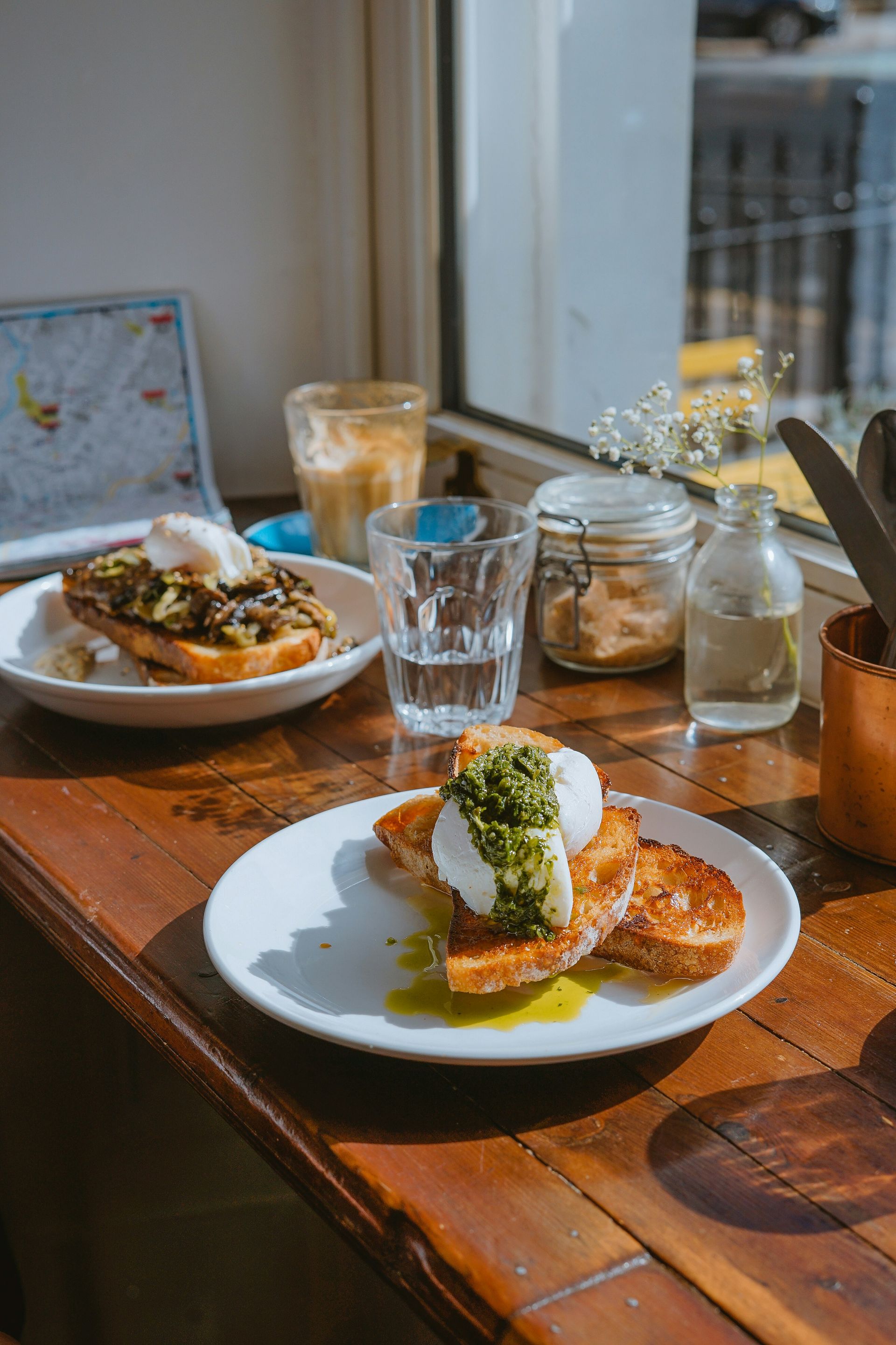 Zwei Teller mit geröstetem Brot, belegt mit pochierten Eiern und grünen Kräutern, auf einem Holztisch in der Nähe eines Fensters, dazu Getränke.