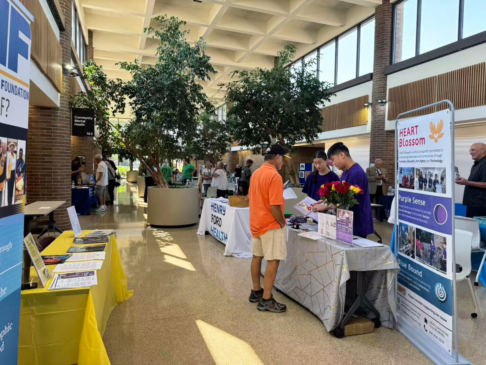 People at tables with information, plants in center. Inside large building.
