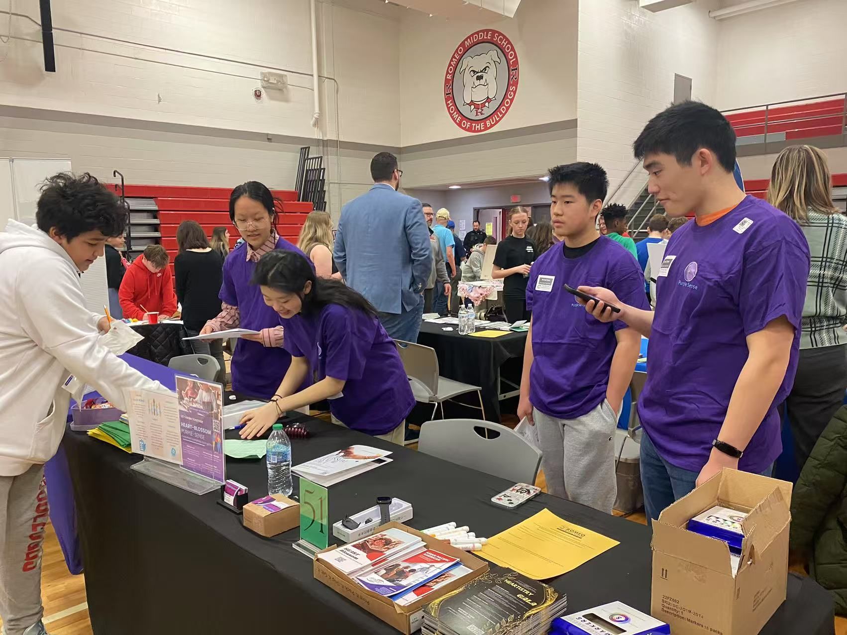 People at a table, possibly a school event. Several wear purple shirts, examining materials. A gymnasium setting.