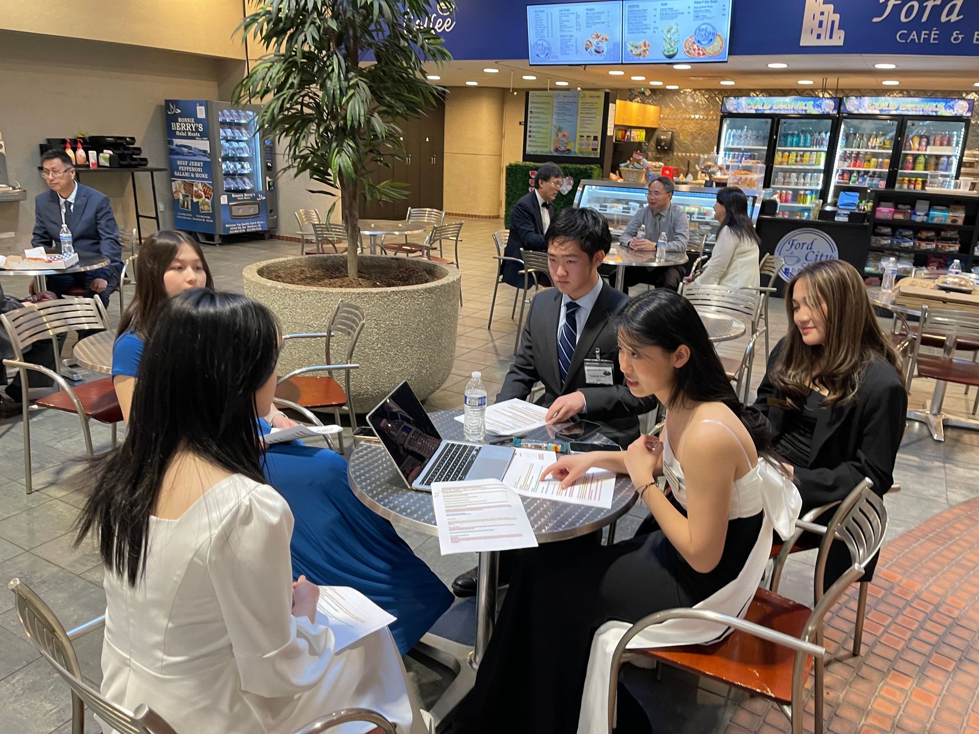 Group of people in formal attire at a cafe table, looking at documents and a laptop.