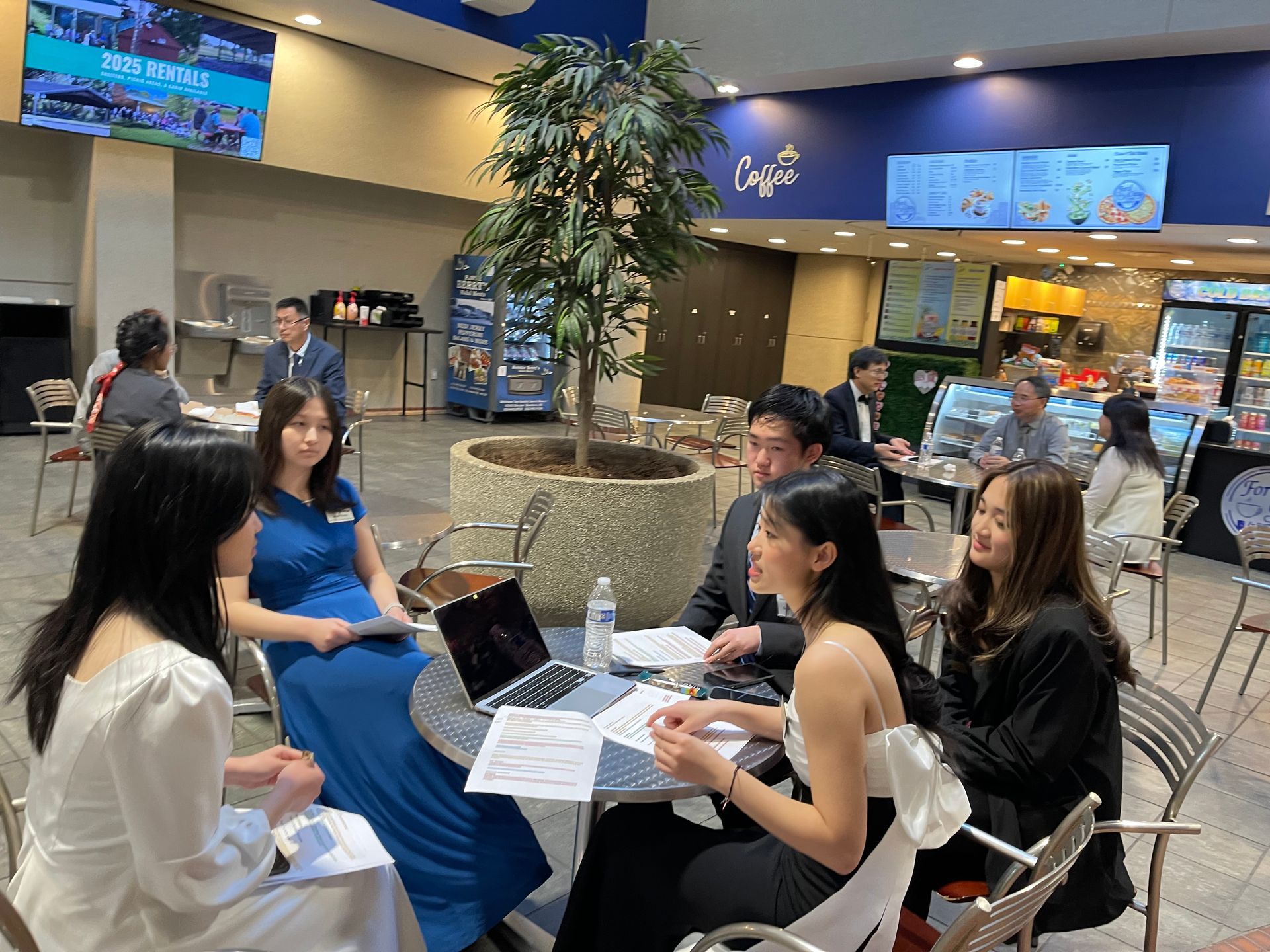 Group of young people in formal wear at a table with a laptop. Indoors, near a cafe.
