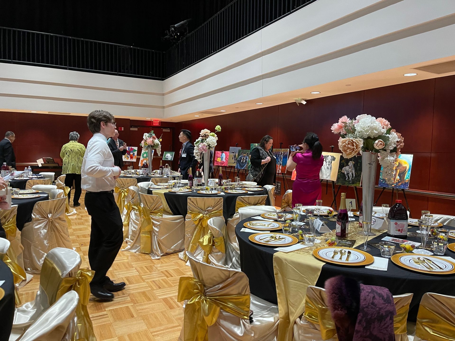 Formal event: round tables with gold accents, paintings on easels, people mingling.