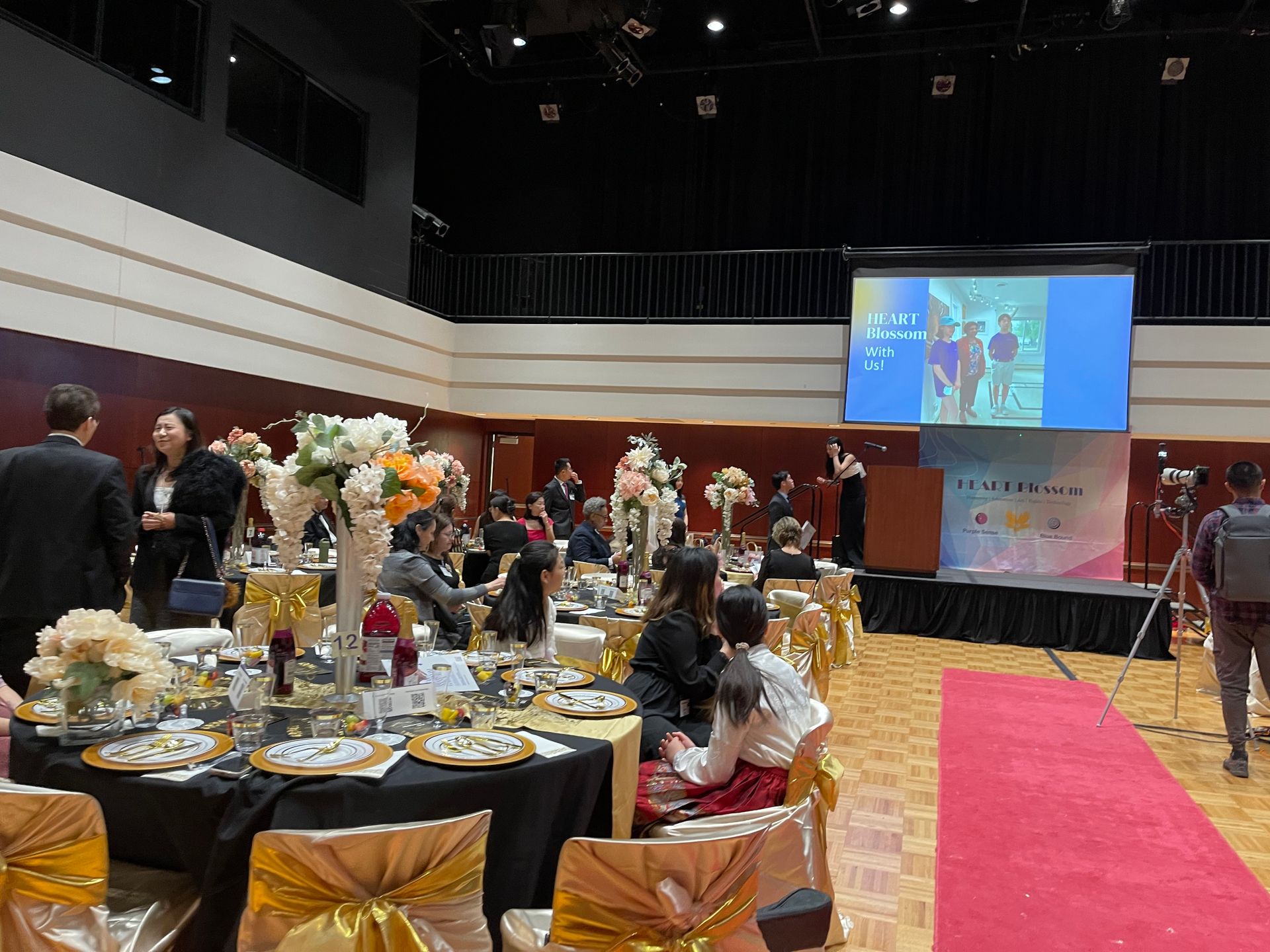 Event in a hall with decorated tables, a stage with a screen, and people attending.