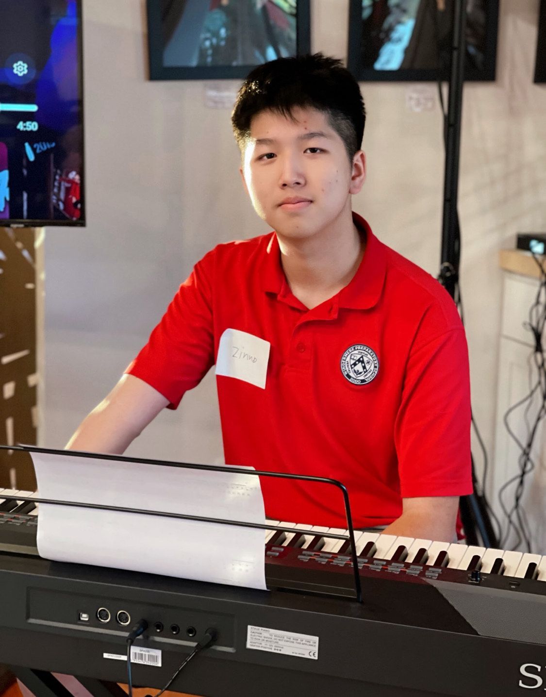 Young man in red shirt at a keyboard, facing camera. Setting appears to be indoors.