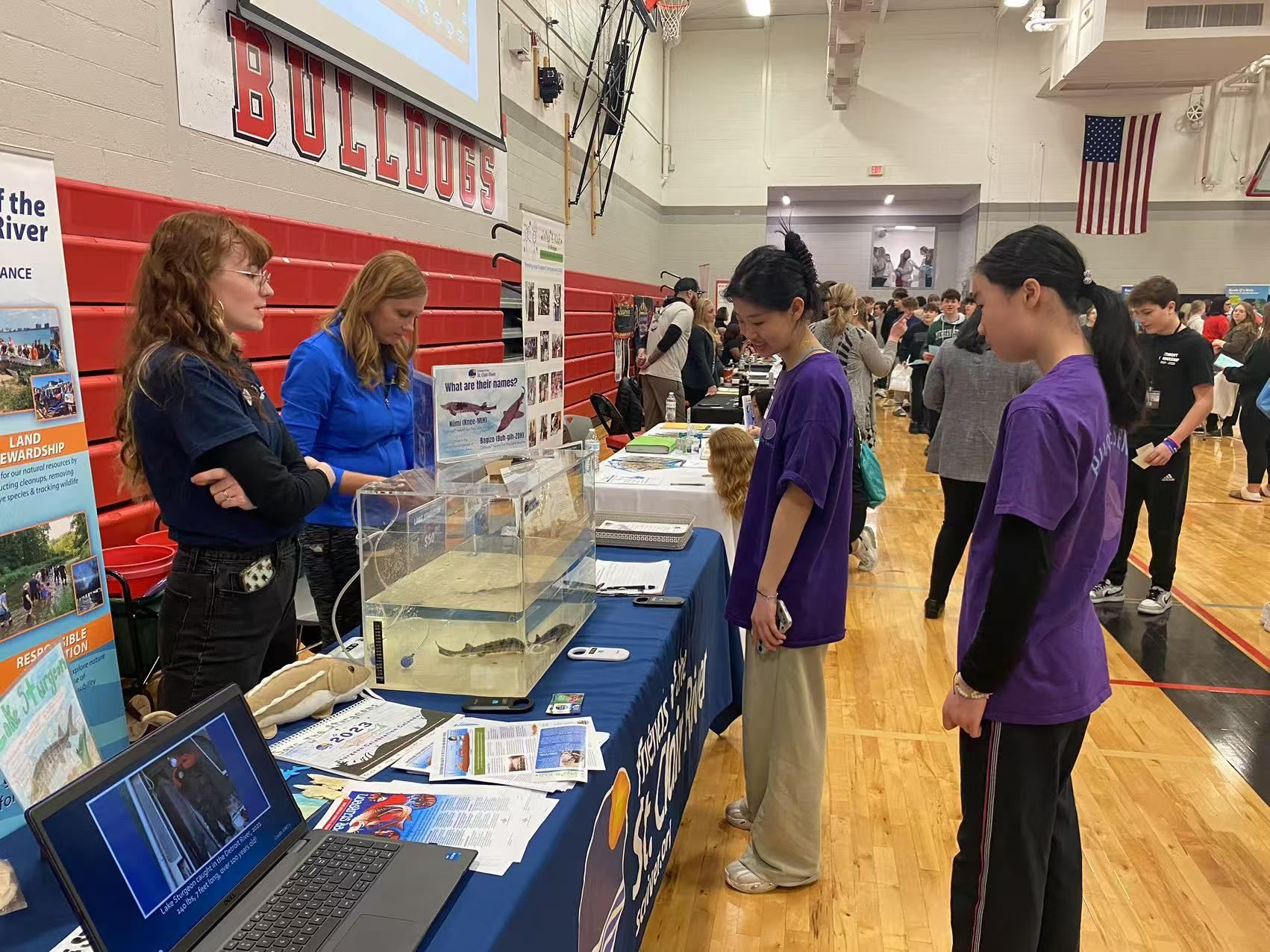 Two women at a table explain information to two girls, with a tank of fish, at a school event.