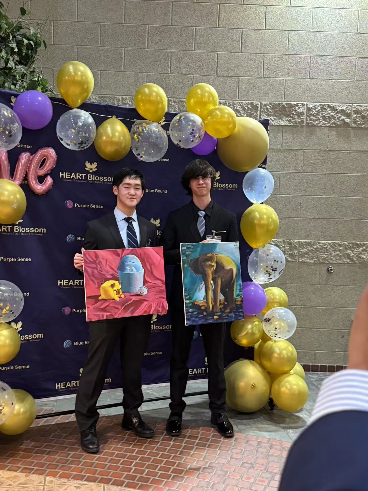 Two young men holding paintings in front of balloons; event setting, suits, smiling.