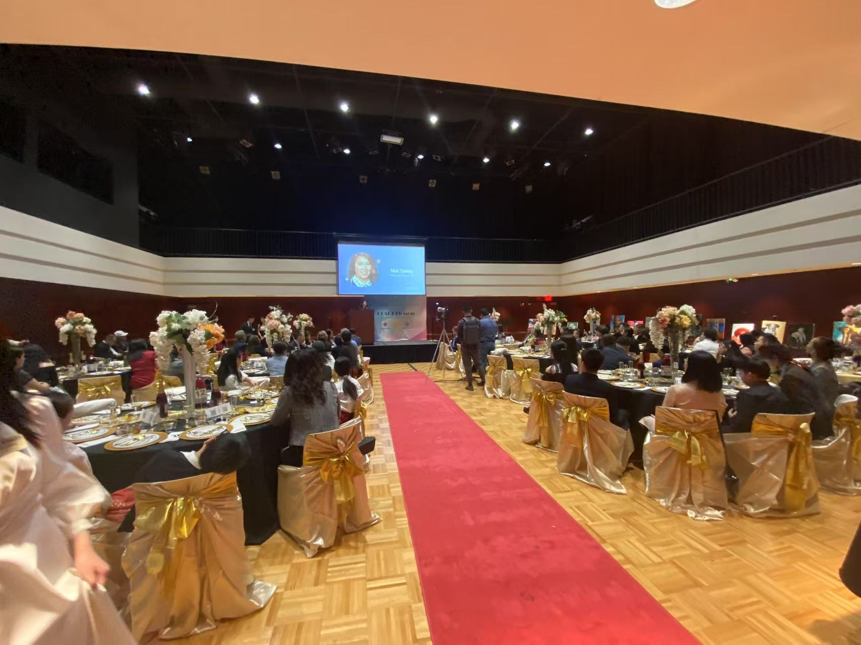 Large banquet hall with tables set for a formal event; red carpet aisle; guests seated; gold accents.