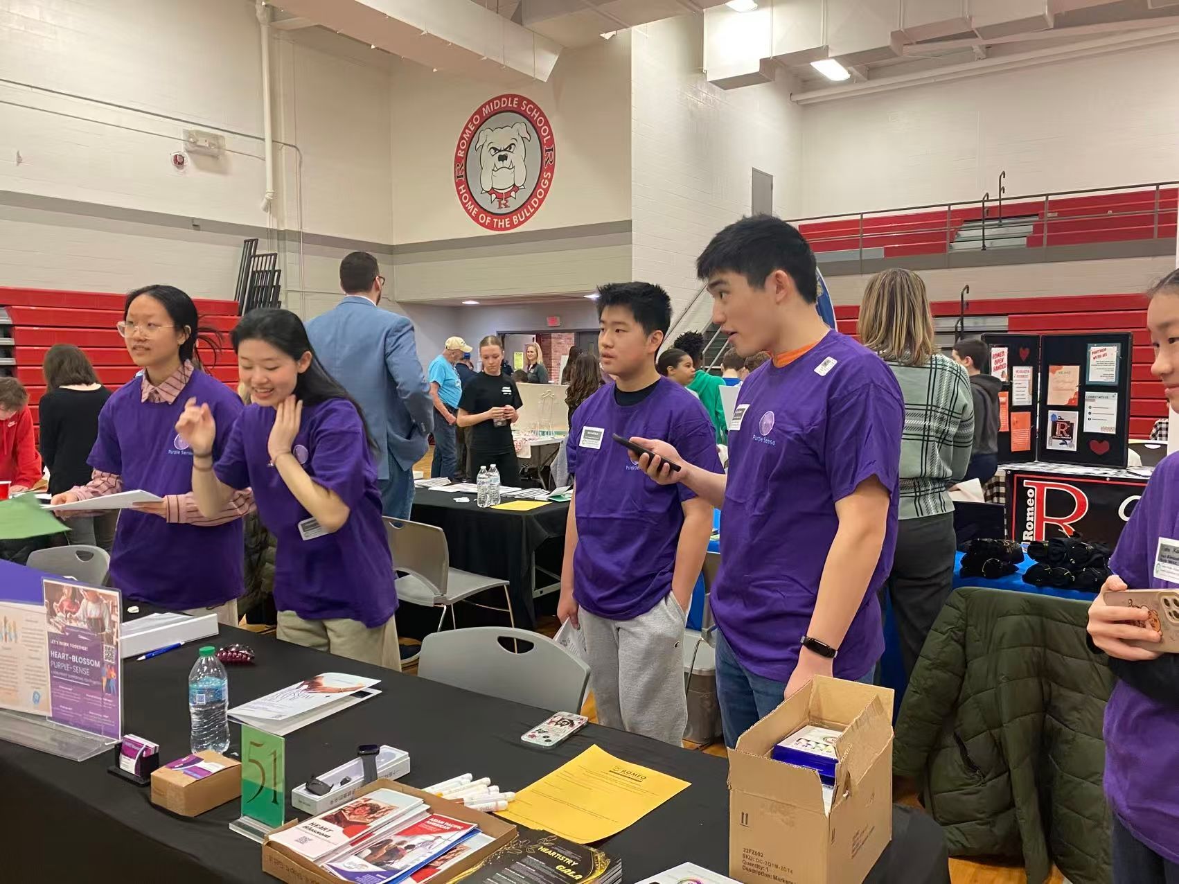 Students in purple shirts at a table, engaging with visitors in a school gymnasium.