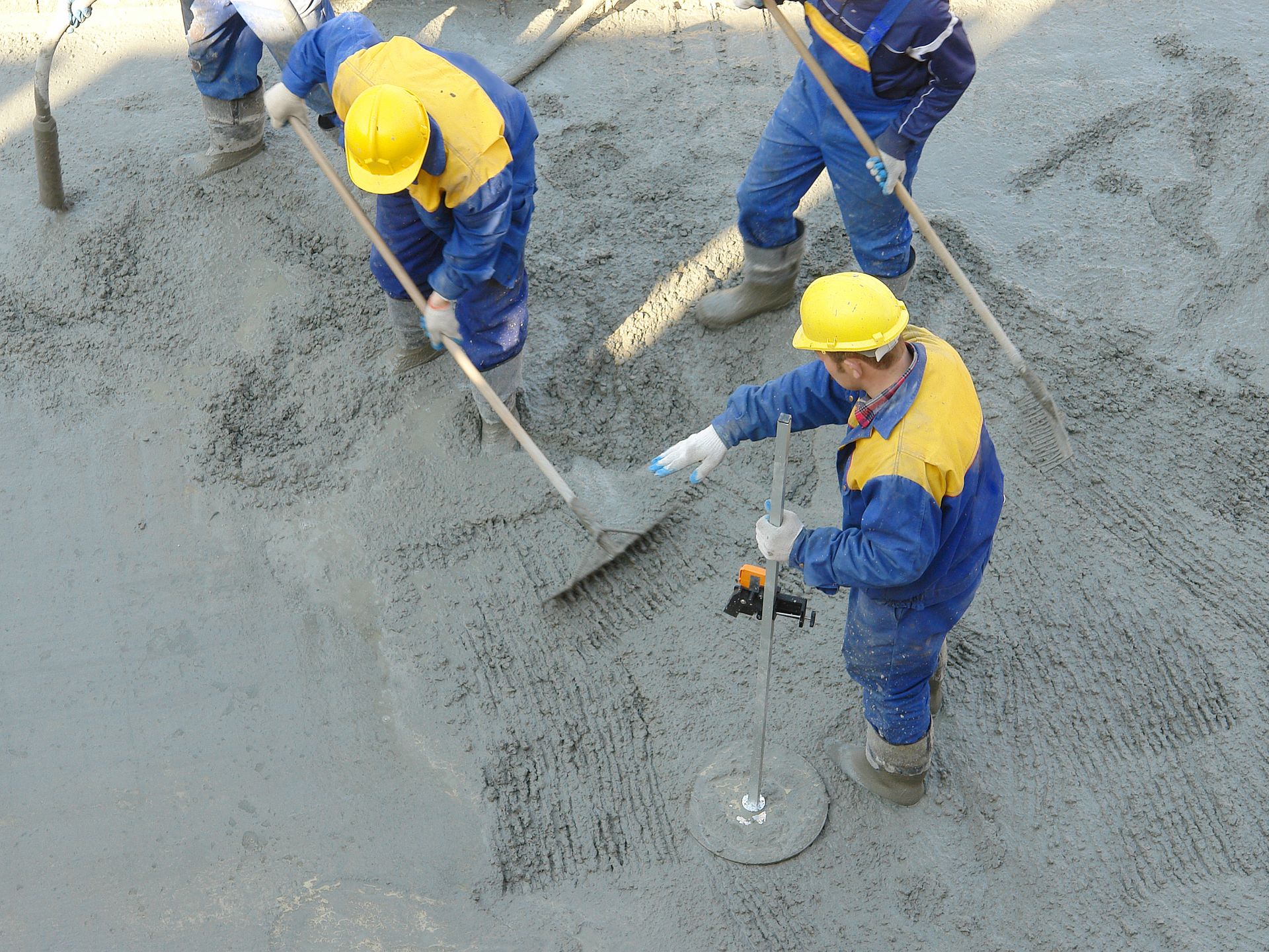 Workers in blue coveralls and yellow helmets leveling wet concrete with hand tools.
