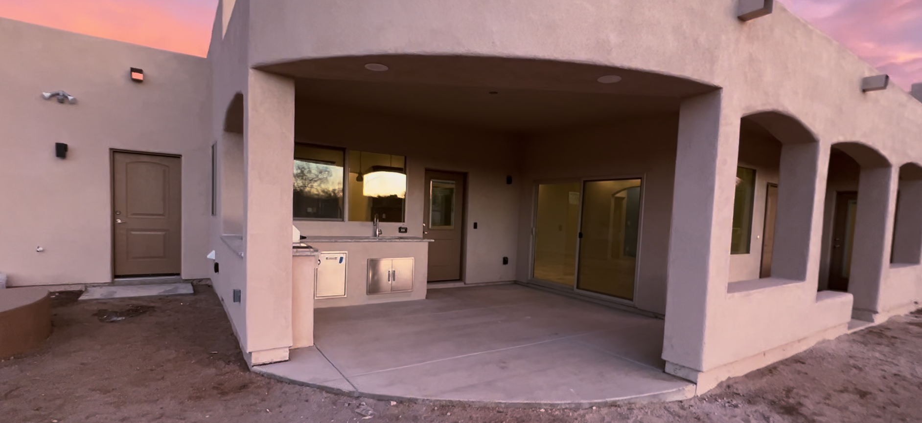 A large house with a patio in front of it and a sunset in the background and an outdoor kitchen