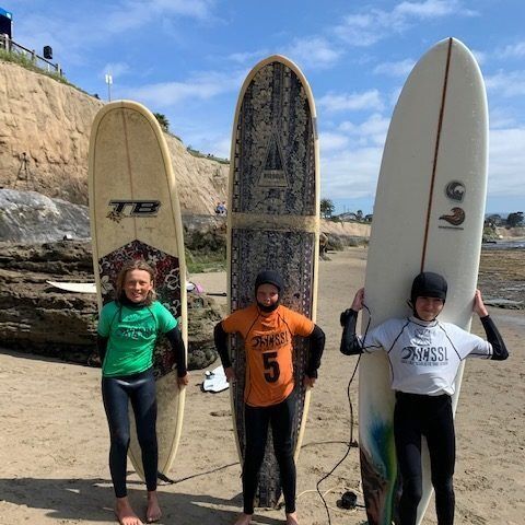 Three surfers holding longboards on a beach beside cliffs and rocky shoreline.