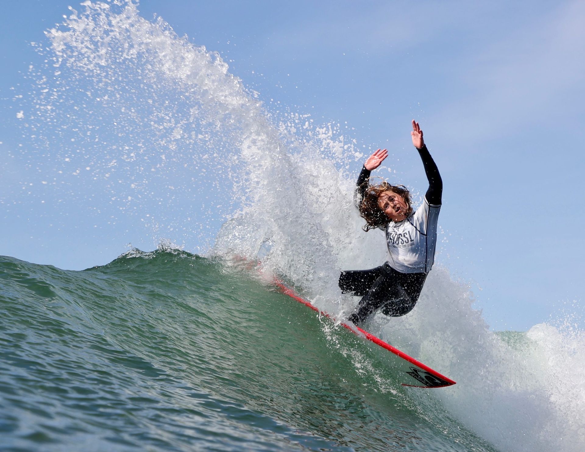 Surfer riding a wave, splashing water with arms raised on a red surfboard under a blue sky