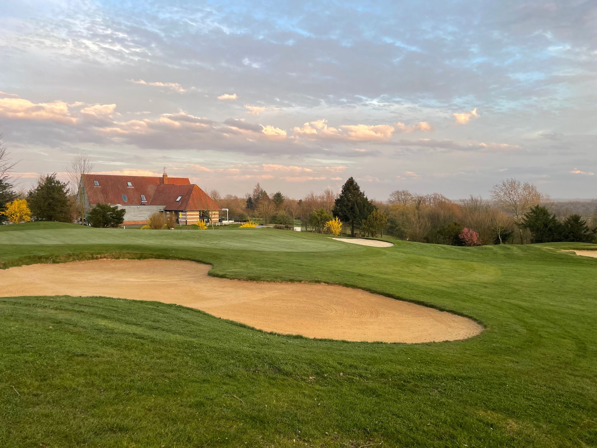 Golf course green with sand bunker under a cloudy sunset sky, houses in the distance