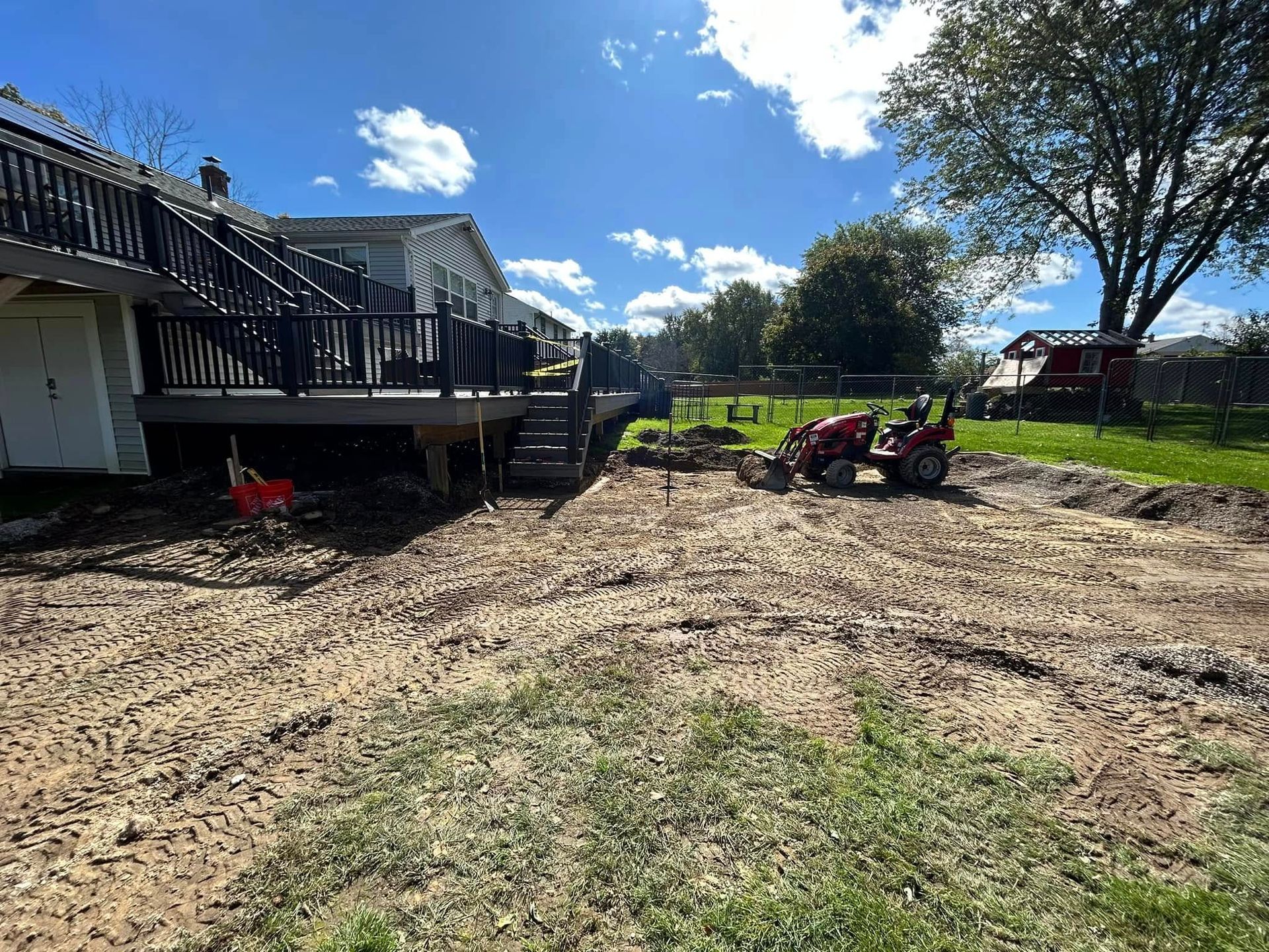 Backyard with muddy ground, a raised deck on the left, and lawn equipment near trees under a blue sky