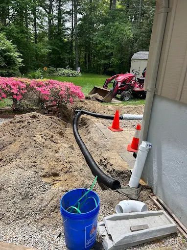 Backyard construction site with a red skid steer, orange cones, blue bucket, and black pipe beside a house