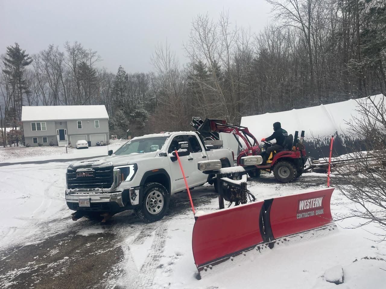 Plow truck and ATV working in snowy driveway beside red snowplow barrier.