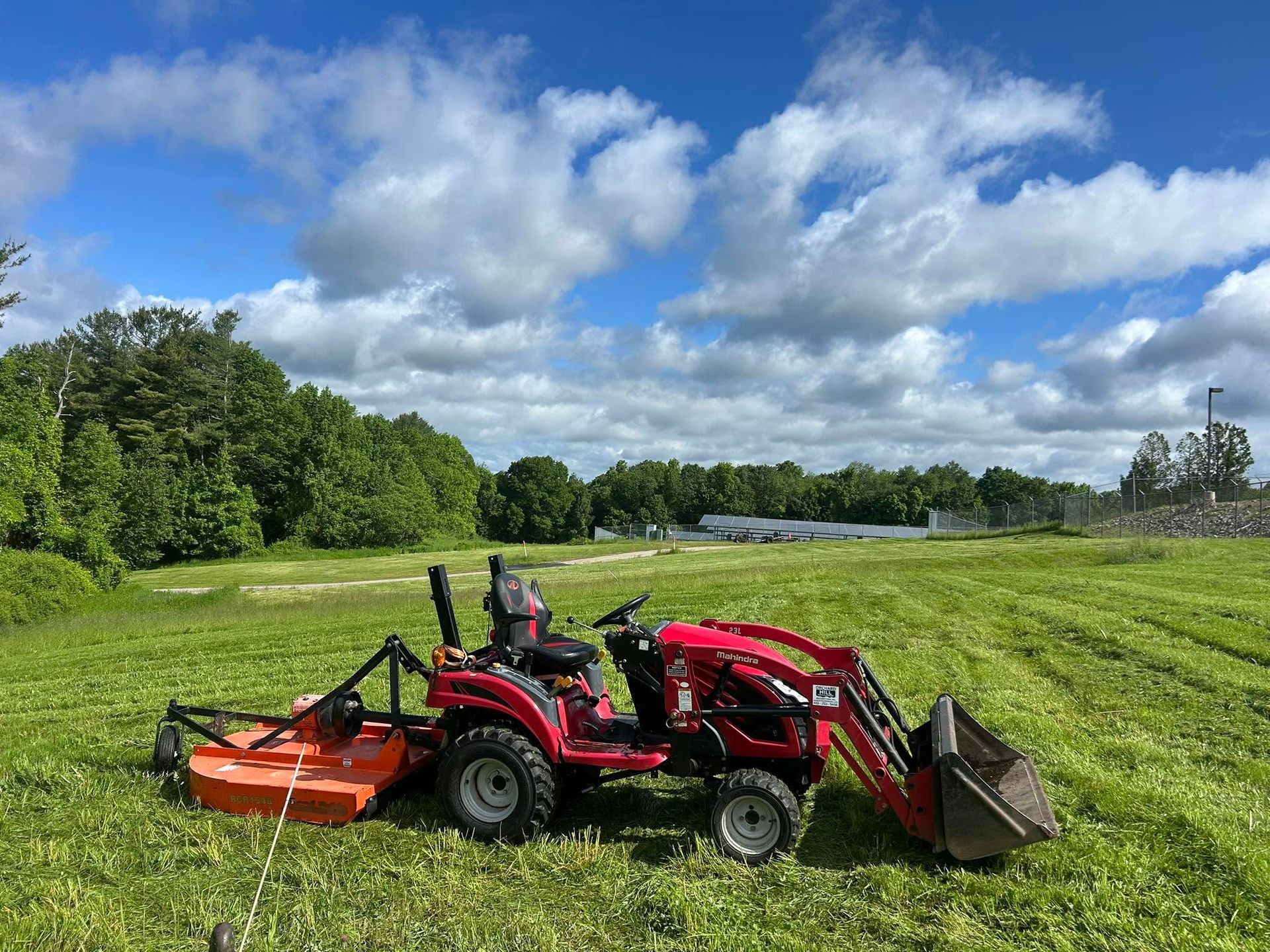 Red tractor mowing grass in a green field under a cloudy blue sky.