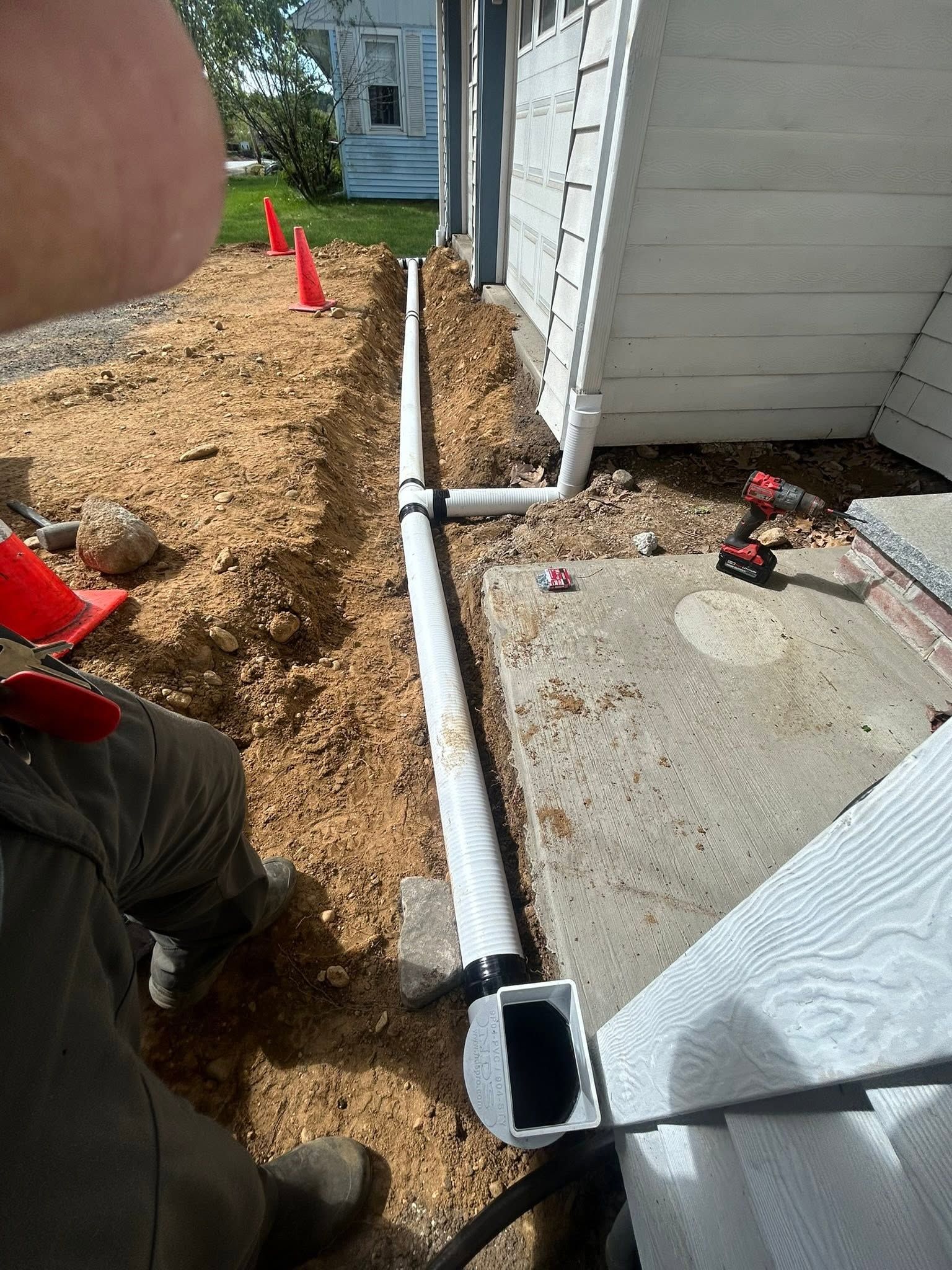 White downspout along house wall beside concrete pad and mulch, with orange cones marking a trench.