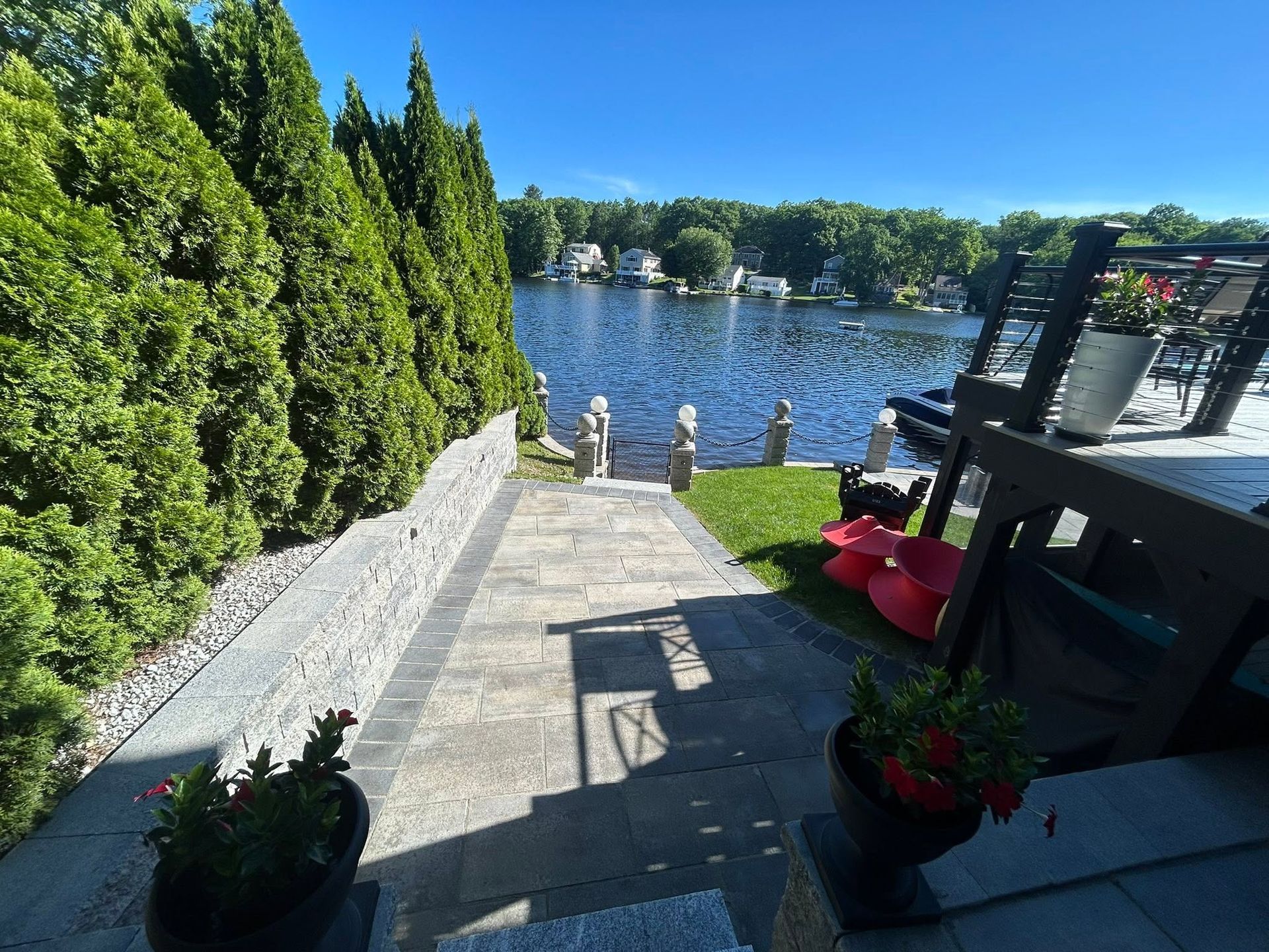 Stone path leading to a blue lake with trees, potted flowers, and a patio chair in the foreground