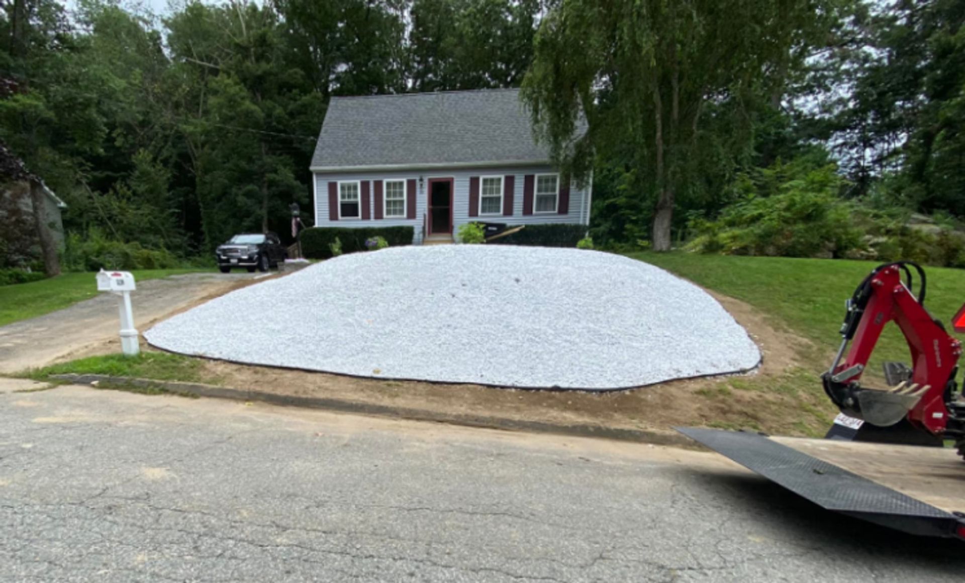 A house front yard covered with white gravel, with a red excavator and a driveway in the foreground.