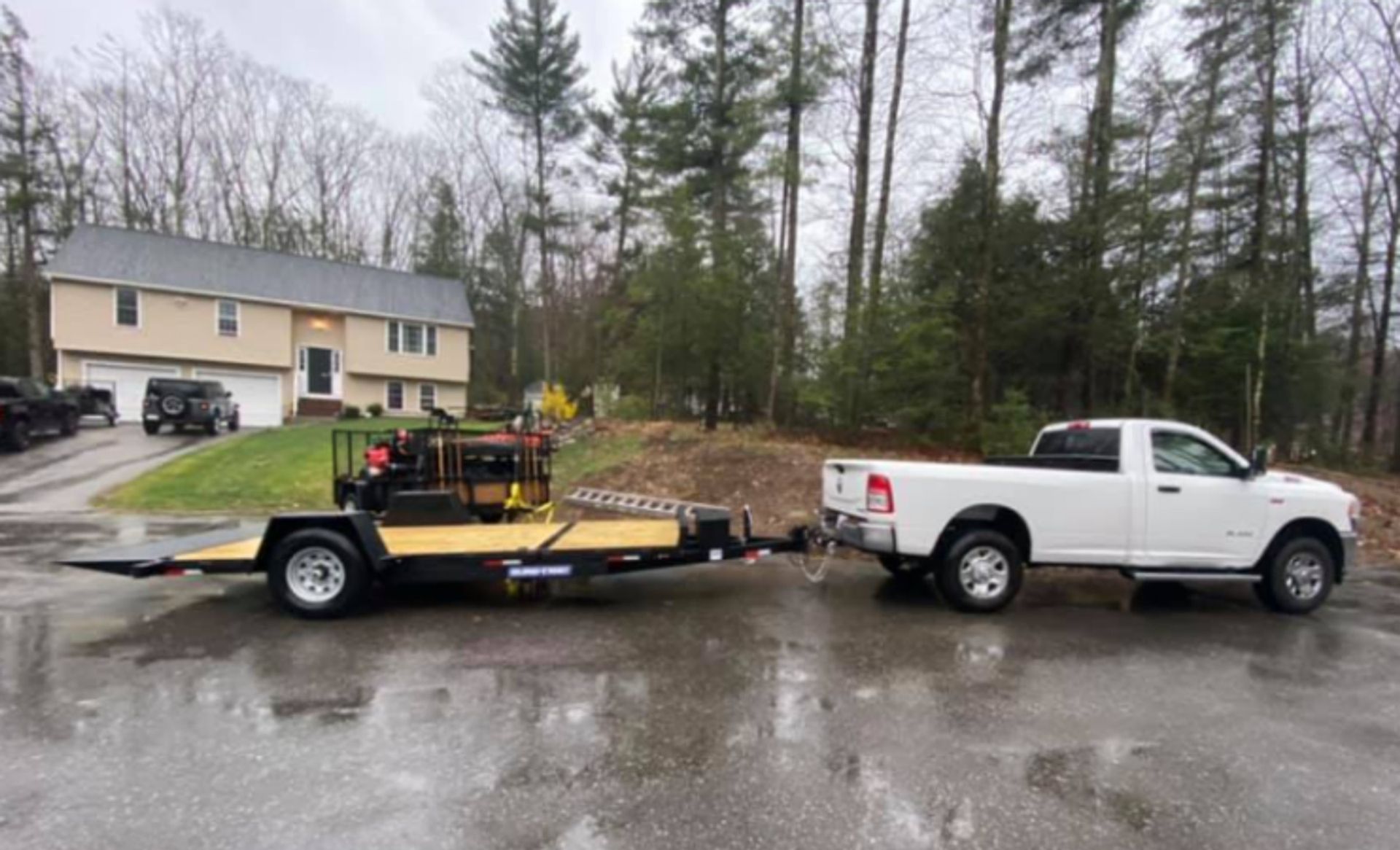White pickup truck towing a flatbed trailer with equipment on a wet driveway near a house and trees