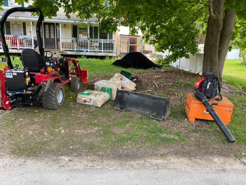 Red lawn tractor, orange leaf blower, and yard supplies on grass under a tree near a house