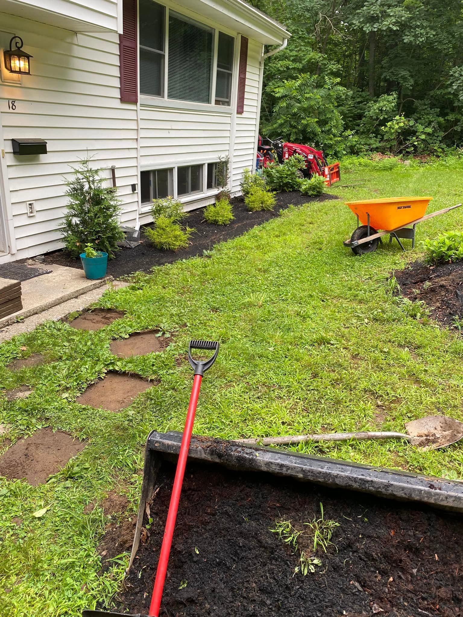 Garden bed edging beside a white house, with a red-handled rake and green grass after rain