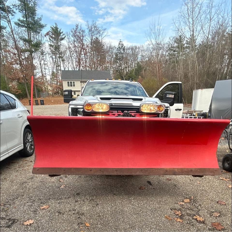 Red snowplow blade mounted on a truck in a driveway, with another car at left and trees in the background