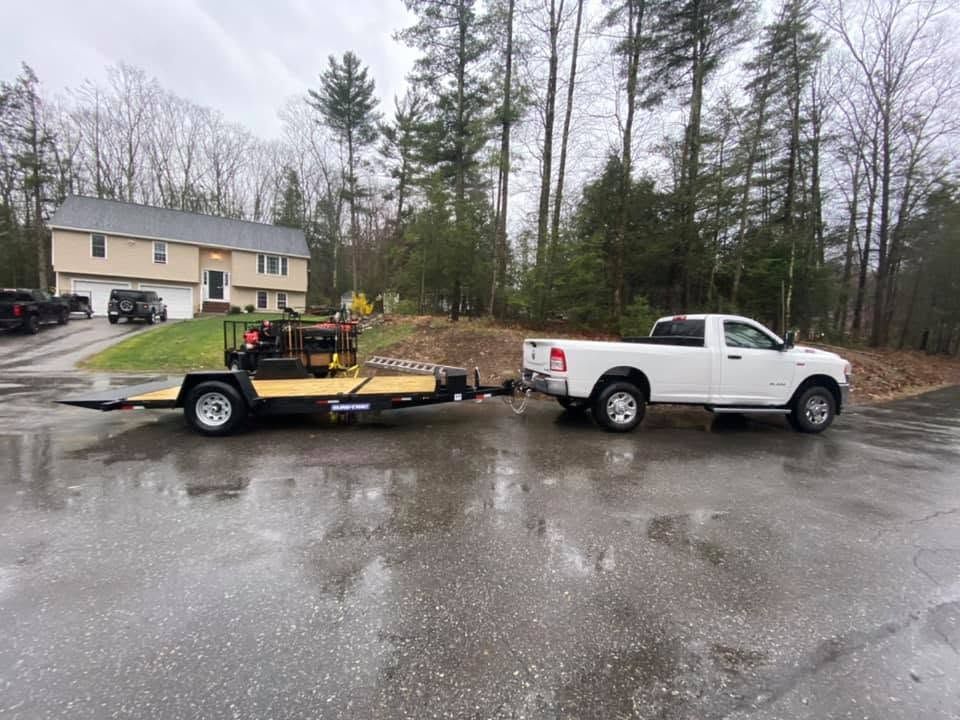 White pickup truck towing a black trailer in a wet parking lot near a house and trees