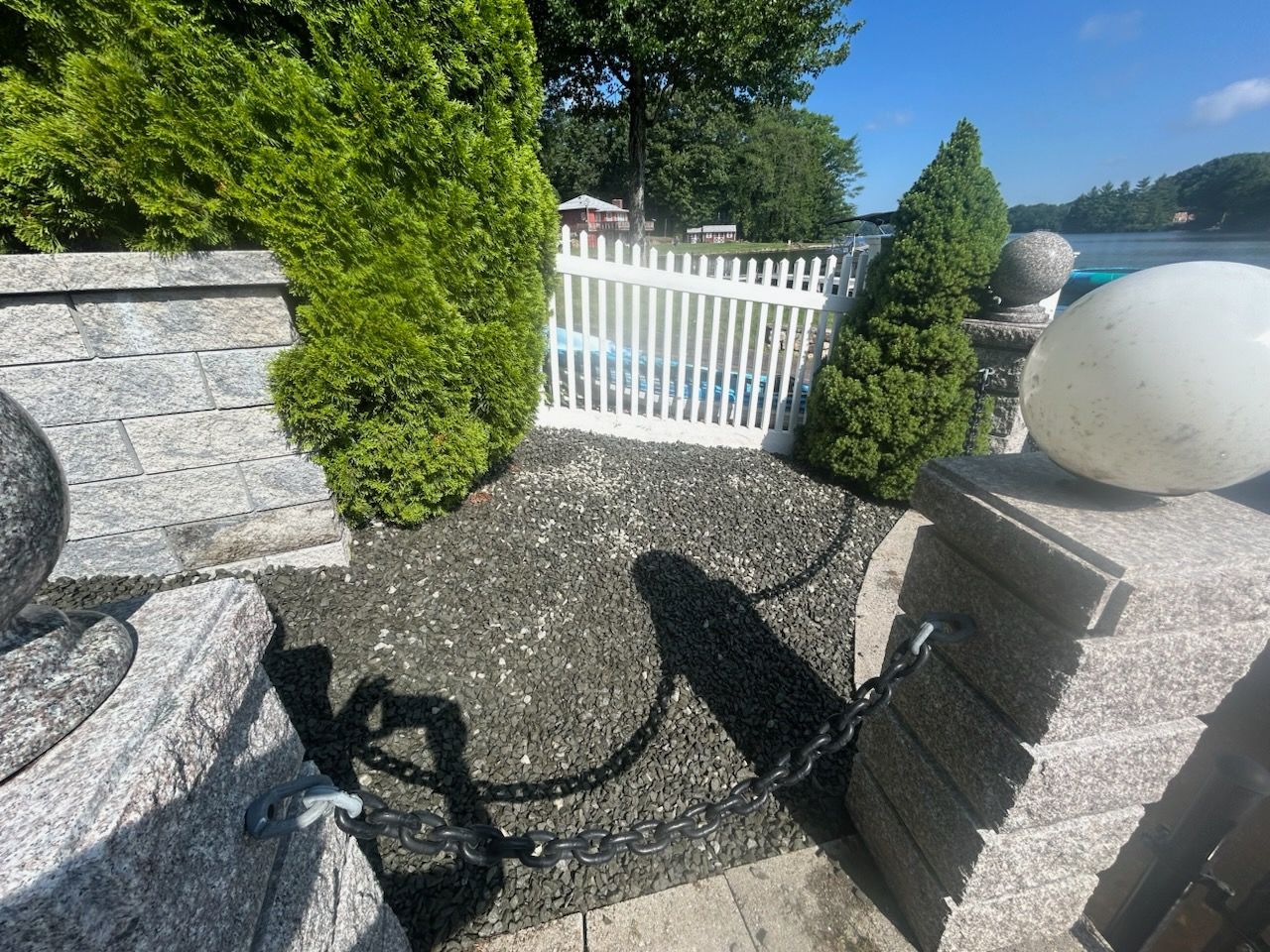Gravel path by stone wall and white gate in a sunny garden, with shrubs and sea view in the distance