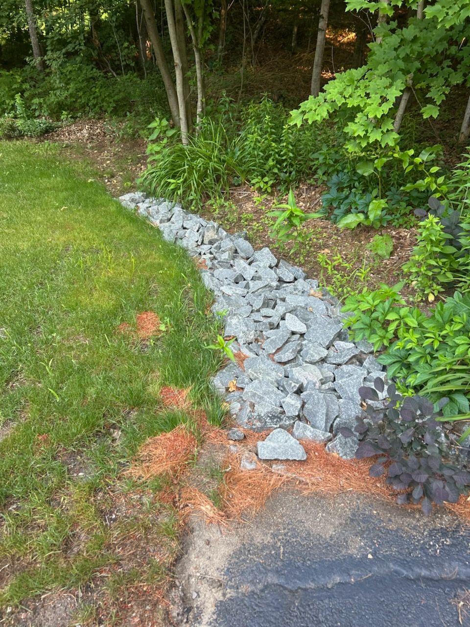 Gray rocks lining a drainage strip beside a grassy yard and wooded edge.