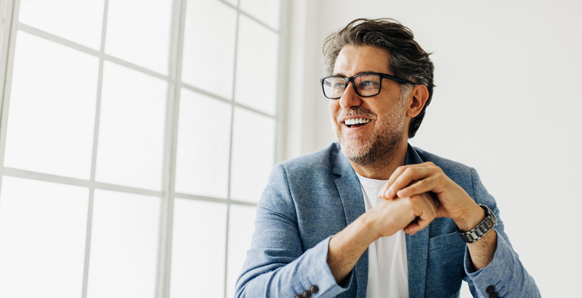 A man wearing glasses is smiling while sitting in front of a window.
