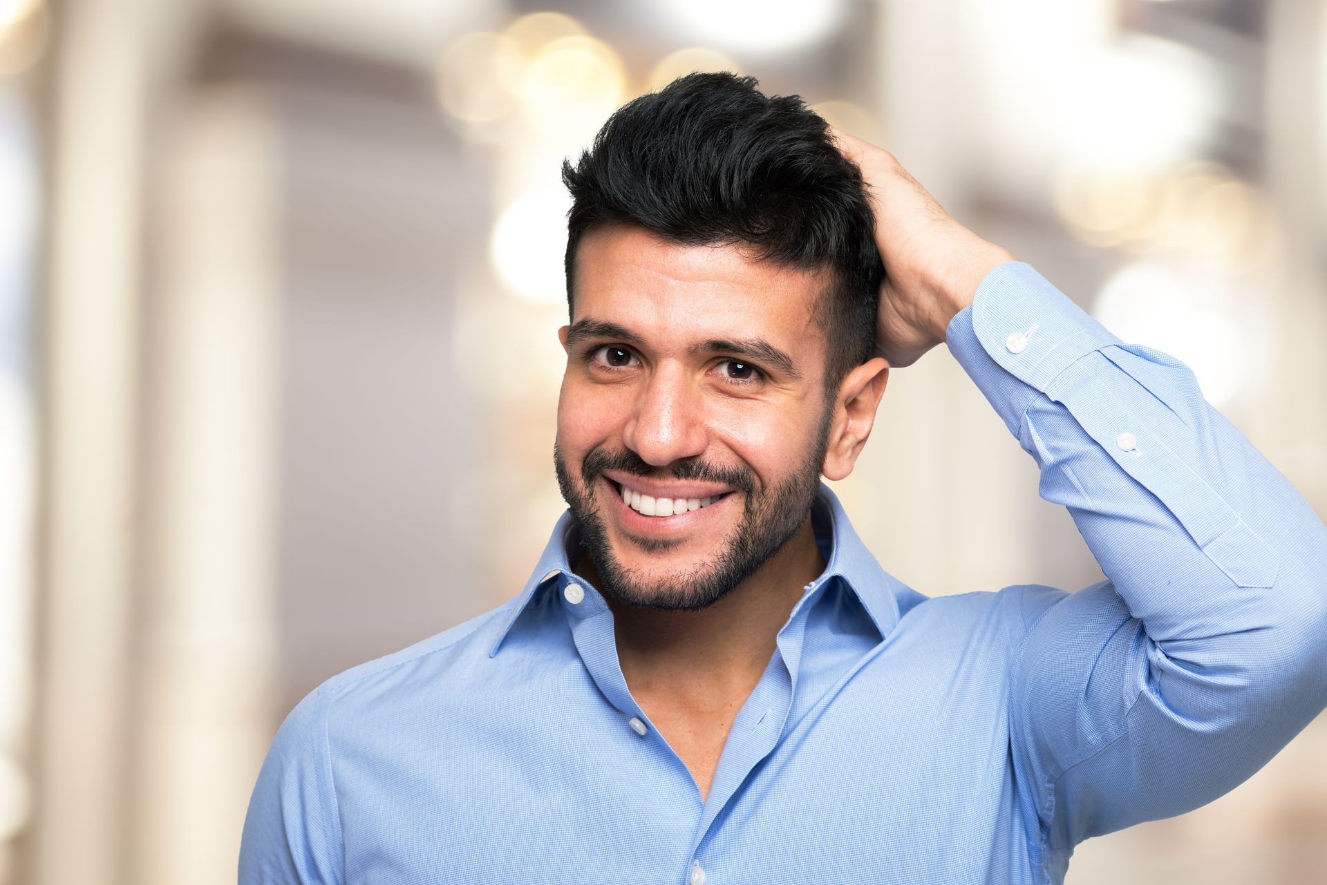A man in a blue shirt is smiling and touching his hair.
