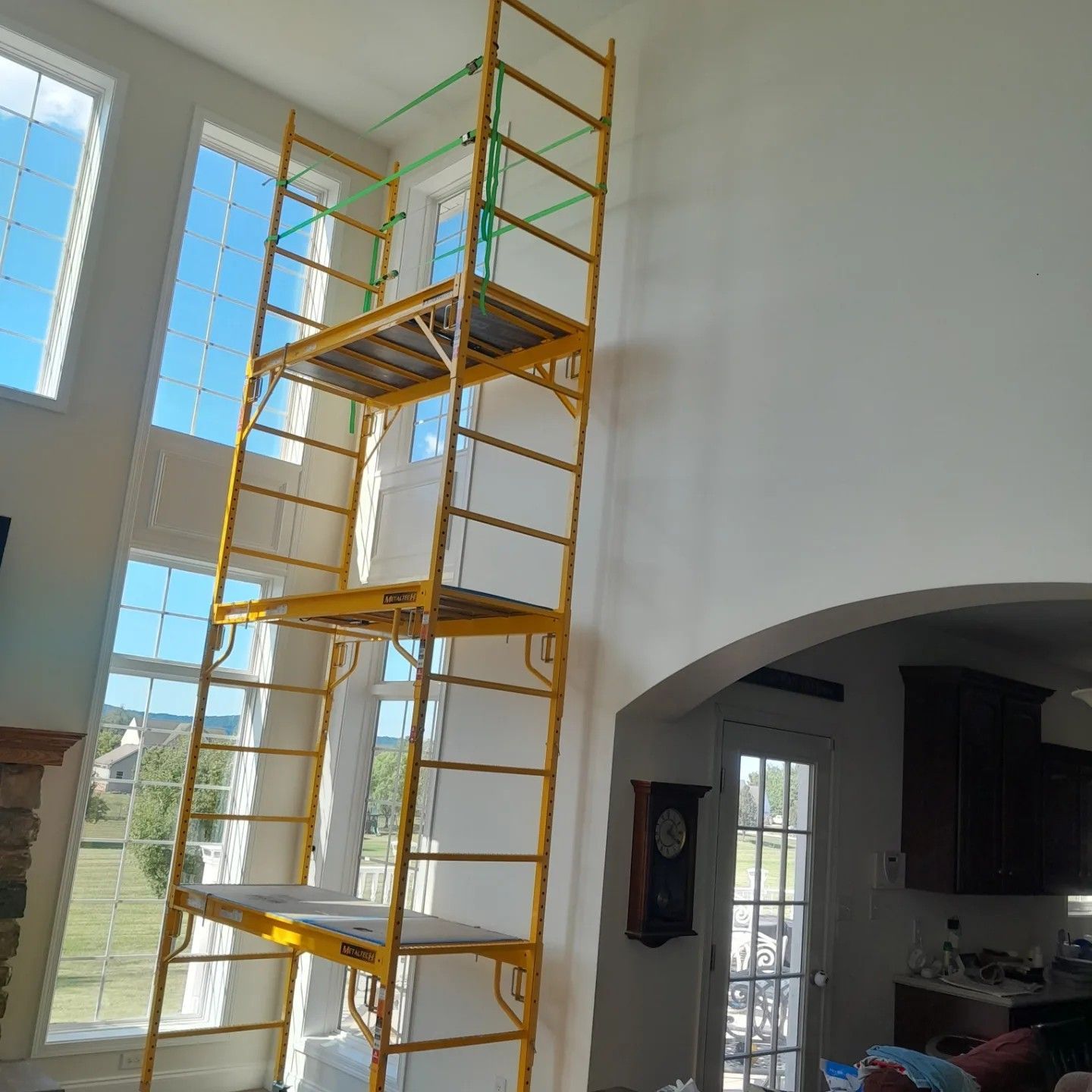 A tall, three-level yellow metal scaffolding tower standing in a high-ceilinged room next to large windows.