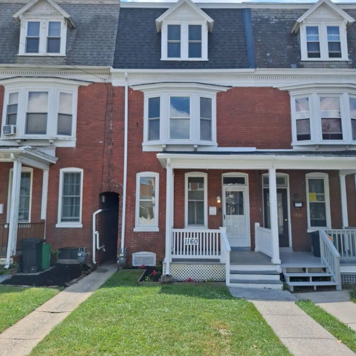 A row of three brick townhouses with dormer windows, porches, and front yards under a bright blue sky.