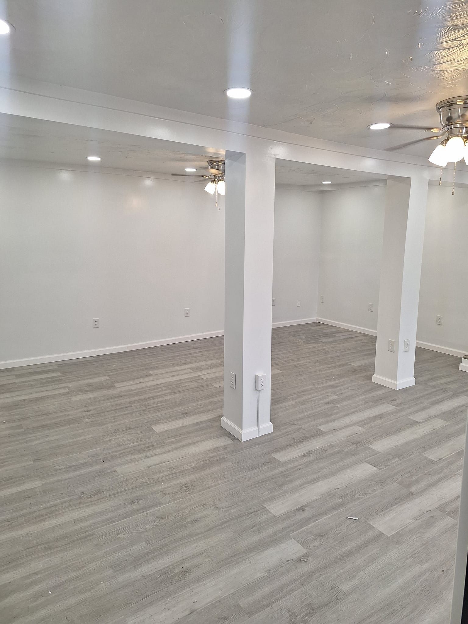 A room with white walls, two white support columns, gray wood-look flooring, and recessed ceiling lights.