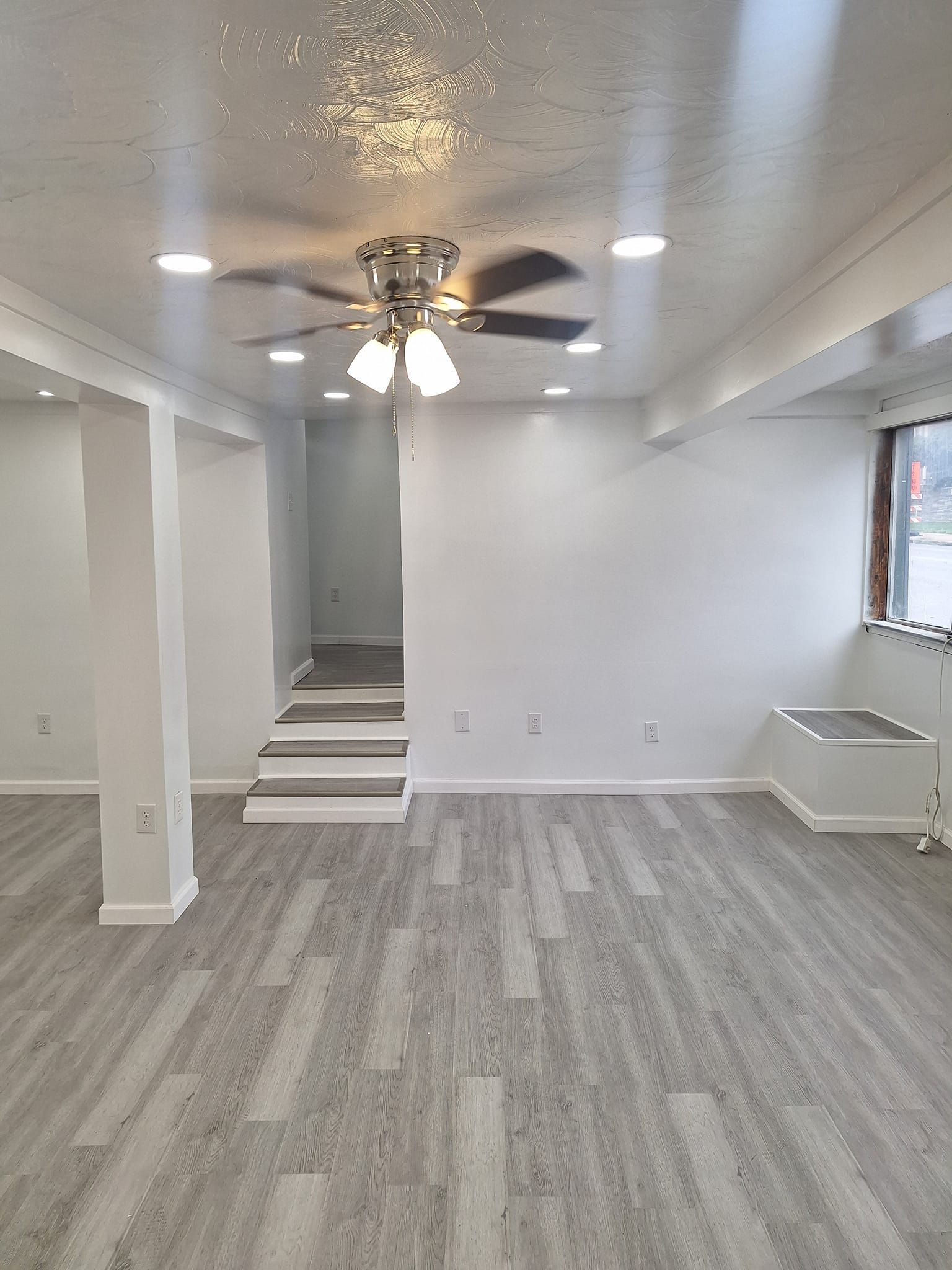 A bright, minimalist room with white walls, grey wood-look flooring, a ceiling fan, and stairs leading to a hallway.