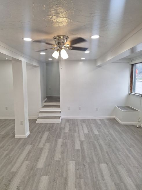 A bright, open-plan room with white walls, recessed lighting, a ceiling fan, gray wood-look flooring, and short stairs.