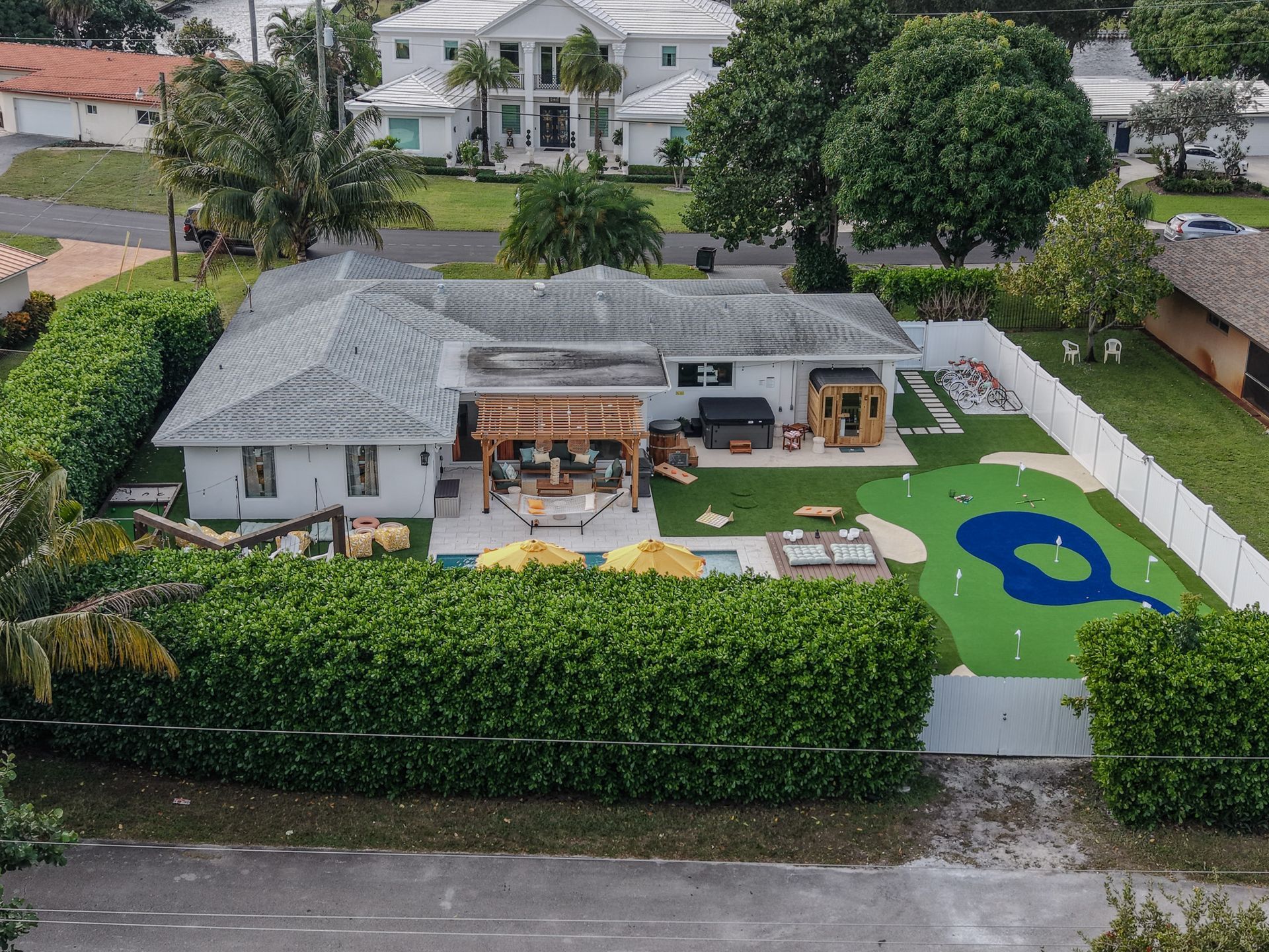 Aerial view of a residential house with a backyard putting green and a patio area enclosed by a white fence and hedges.