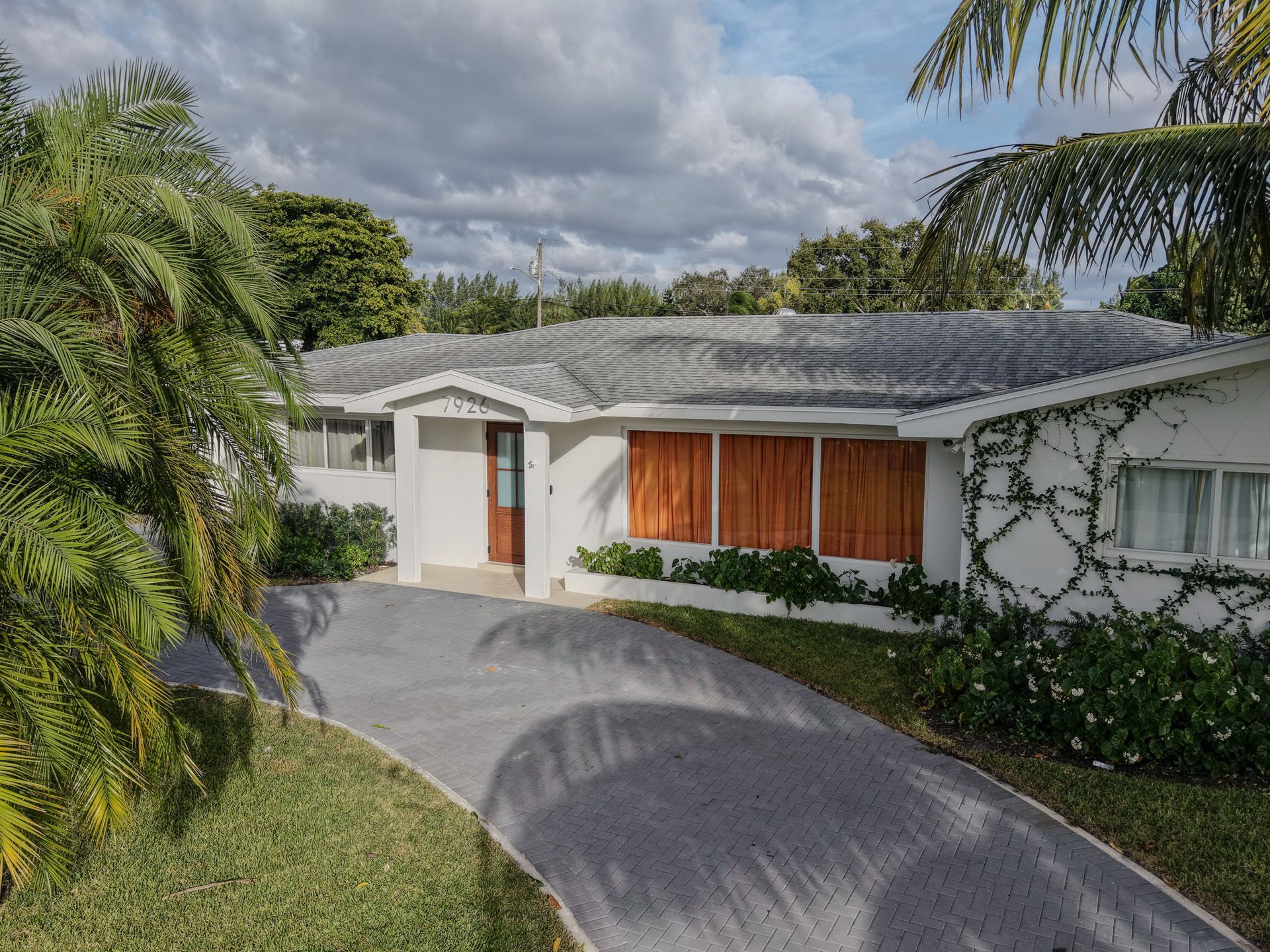 White house with brown shutters, gray driveway, and lush greenery under a cloudy sky.