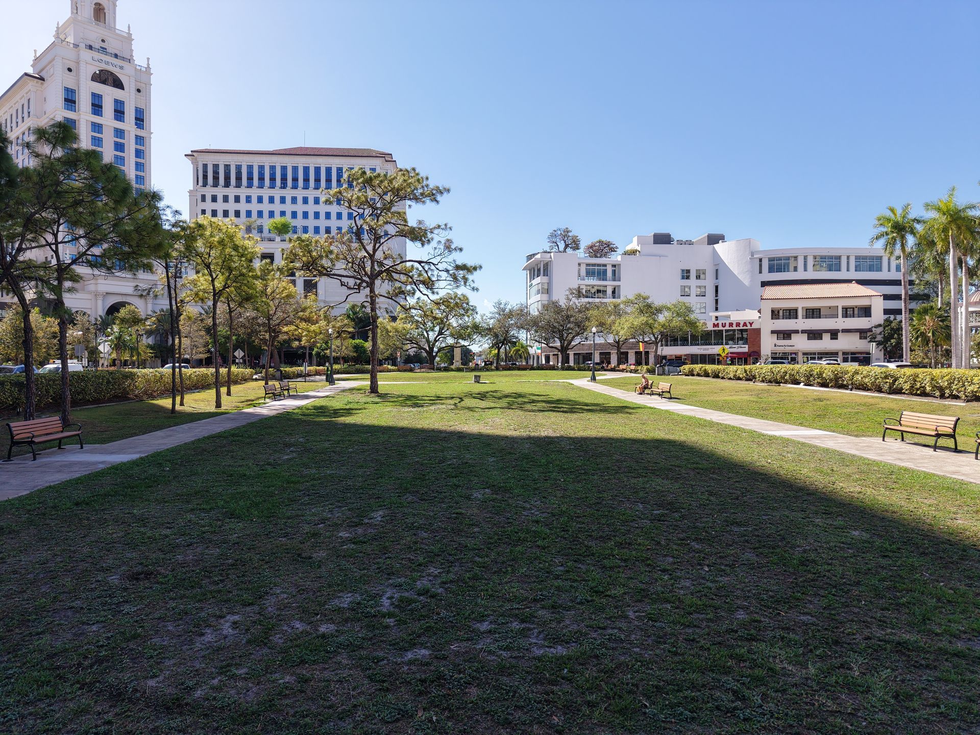 A park area with a grassy lawn surrounded by trees, paved walkways, benches, and city buildings under a clear blue sky.