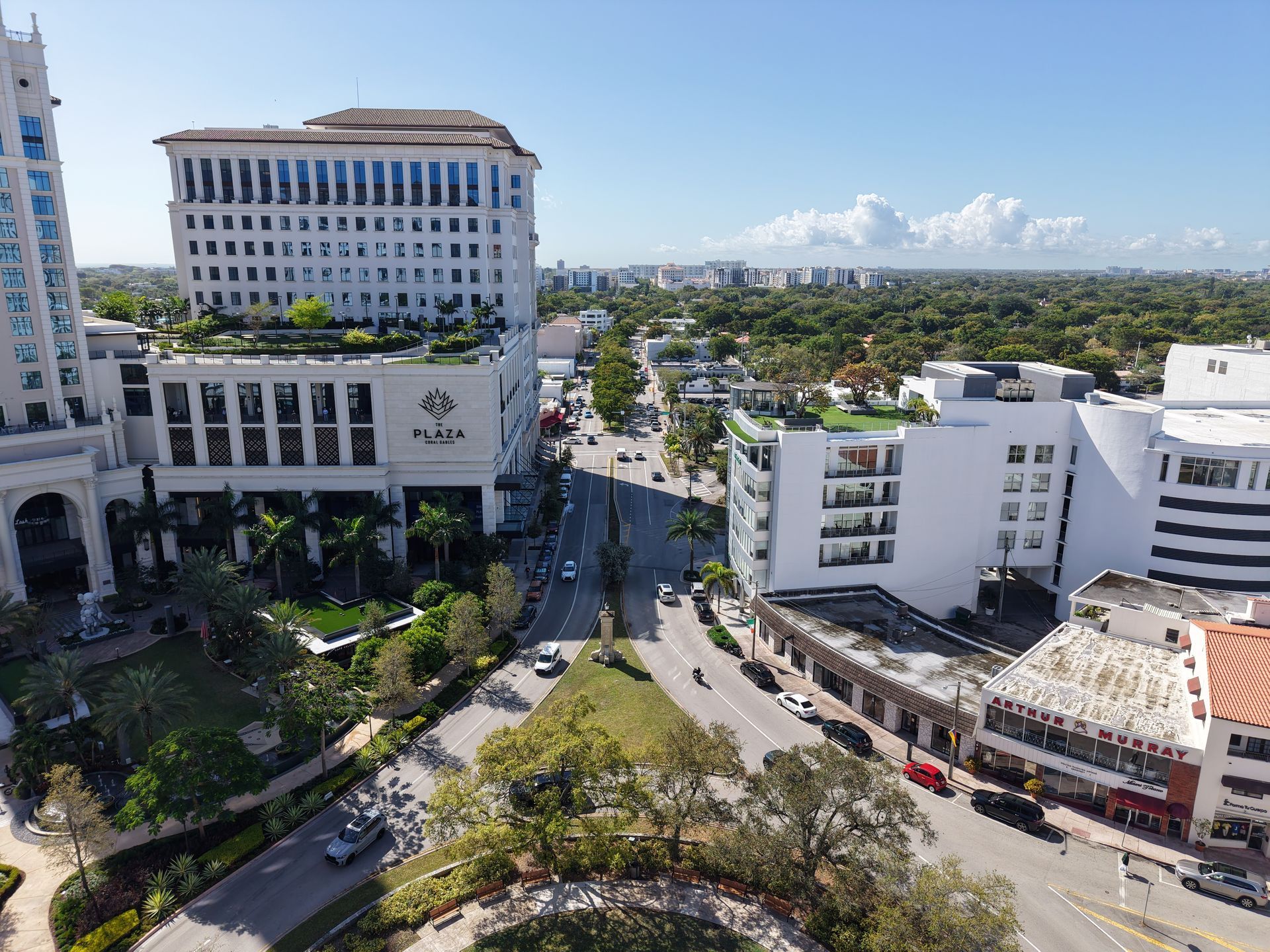 An aerial view of a plaza with a large white hotel, surrounding commercial buildings, streets, and green landscaping.