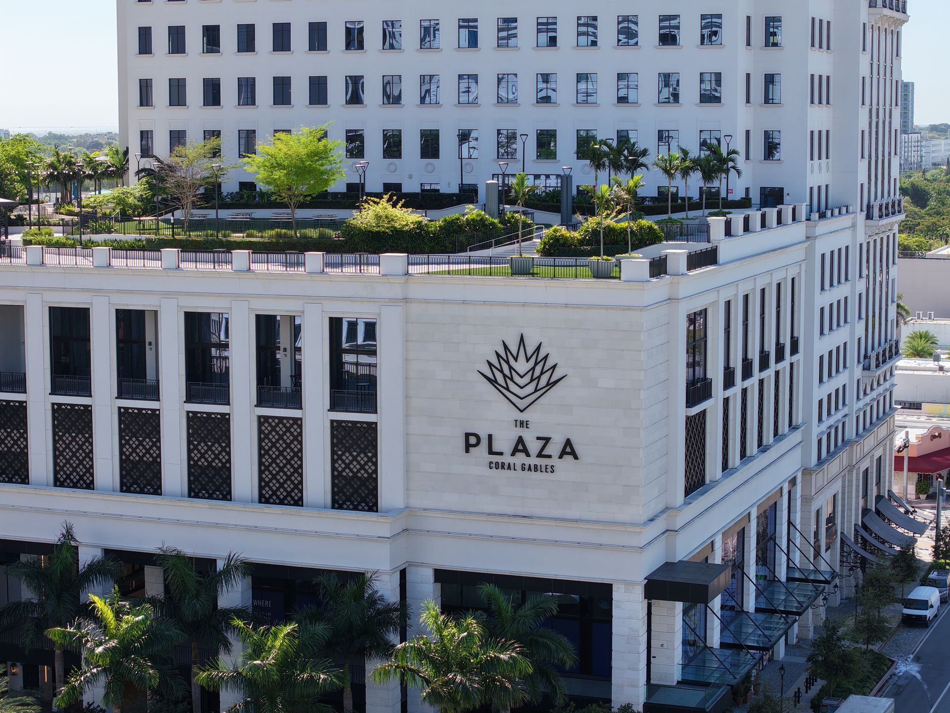 A high-angle view of the cream-colored Plaza Coral Gables building with a landscaped rooftop terrace and palm trees.