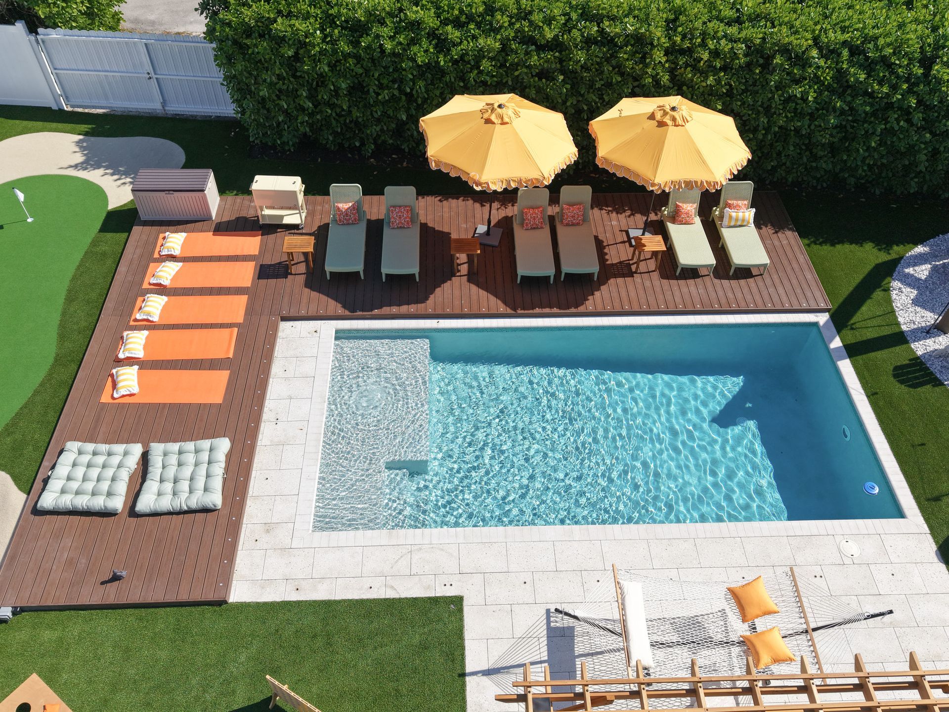 High-angle view of a pool area with orange lounge chairs, two yellow umbrellas on a deck, and light-colored stone patio.
