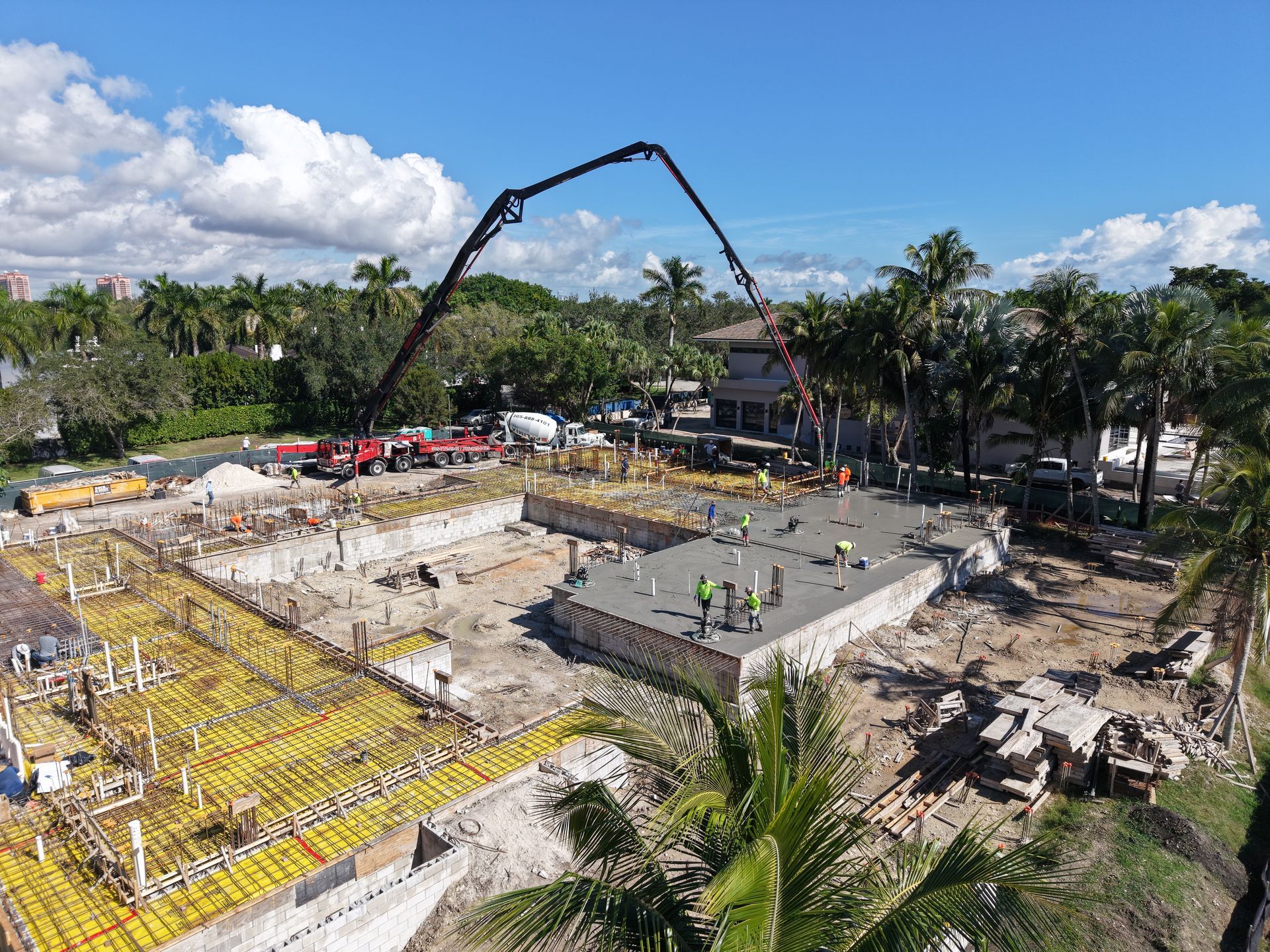 An aerial view shows a construction site with workers pouring concrete using a crane arm over a foundation.
